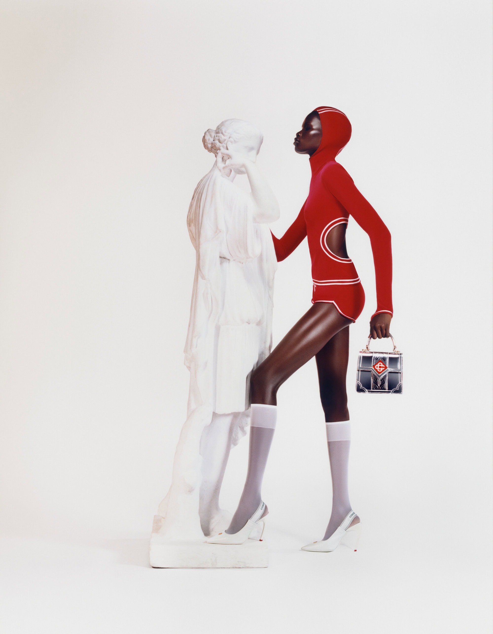 Black model wearing a red hooded bodysuit and white socks standing beside a white classical statue against a seamless background