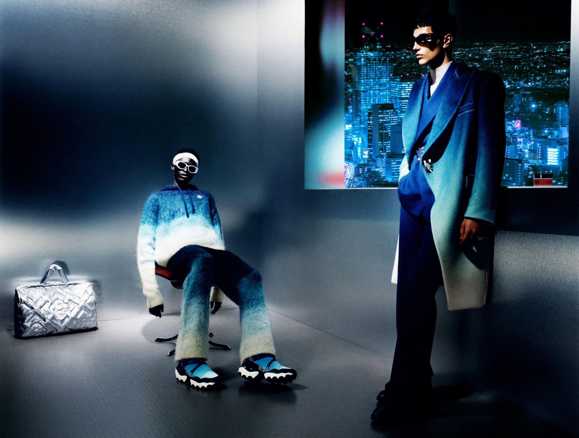 Two models in Casablanca Fall 2024 ad campaign, one seated wearing gradient knitwear, one standing in blue coat before cityscape projection.