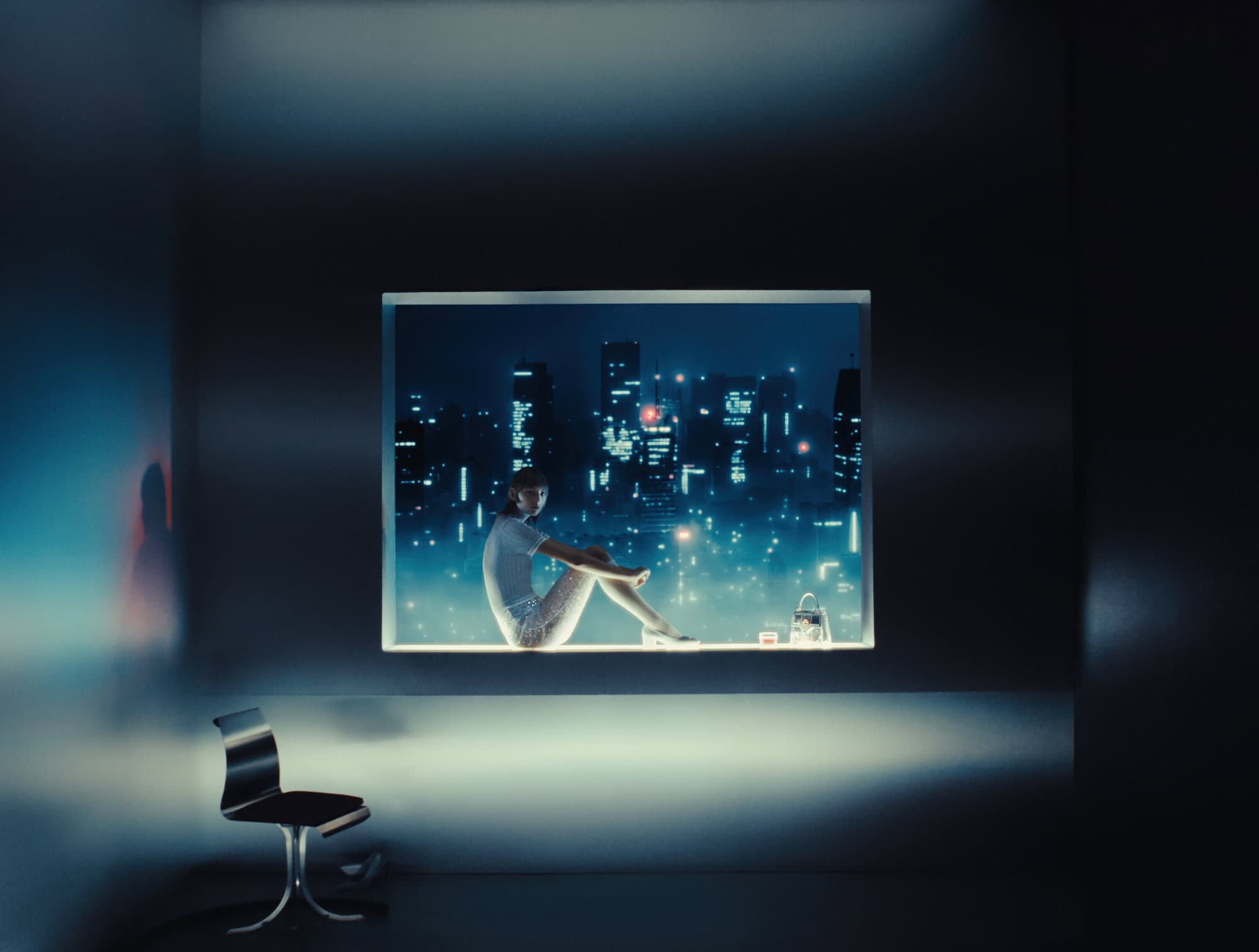 Model seated inside a lit rectangular window aperture against a moody night cityscape backdrop with a modern chair in the foreground.