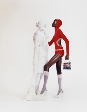 Black model wearing a red hooded bodysuit and white socks standing beside a white classical statue against a seamless background