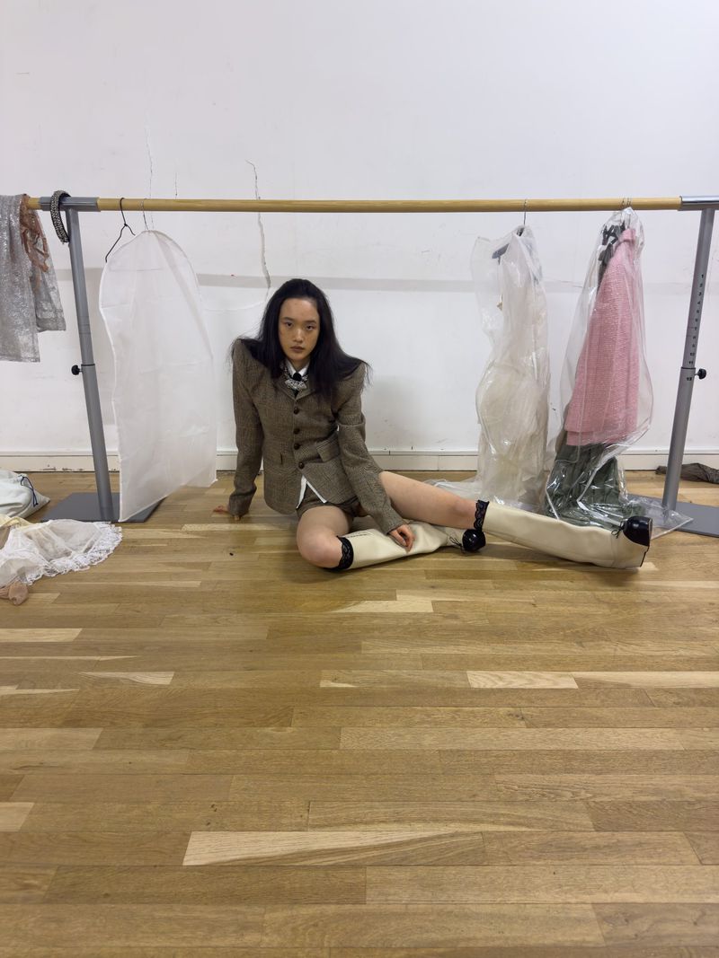 A woman in a vintage suit sitting on a wooden floor with garment bags hanging in the background in a studio setting.