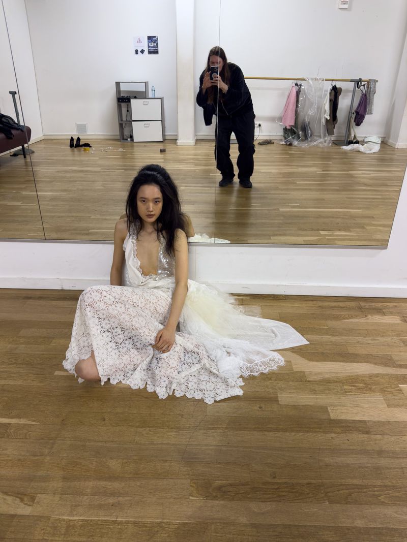 Fashion shoot featuring a woman in a white lace dress in a studio with a photographer visible in a mirror, capturing behind-the-scenes content.