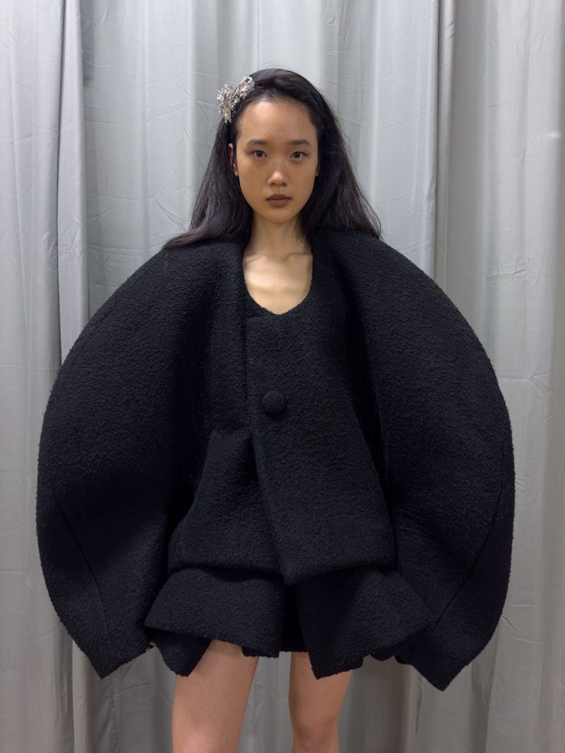 An Asian female fashion model poses indoors in a unique black dress with ruffles, complemented by a sparkly headband, against a grey curtain backdrop.