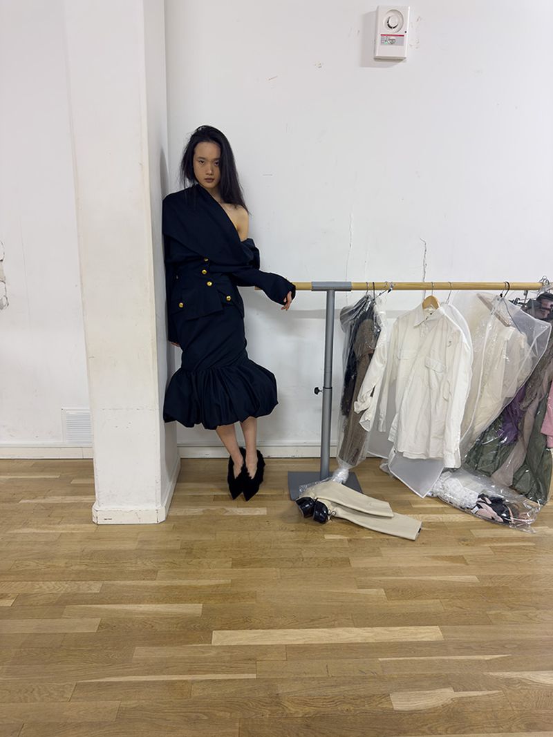 A slender woman poses in a chic black blazer dress with golden buttons, set against a minimalist backdrop with hanging garments.