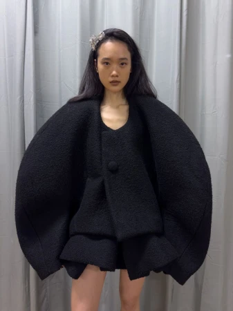 Model wearing a black structured coat with oversized shoulders against a grey curtain background