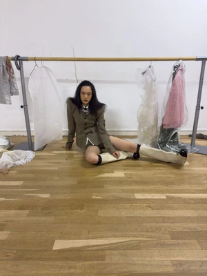 Model seated on a wooden floor wearing a brown patterned suit jacket and white knee-high boots in a backstage dressing room
