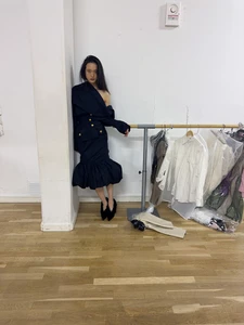 Model wearing dark asymmetric dress leaning against a white pillar next to a clothing rack