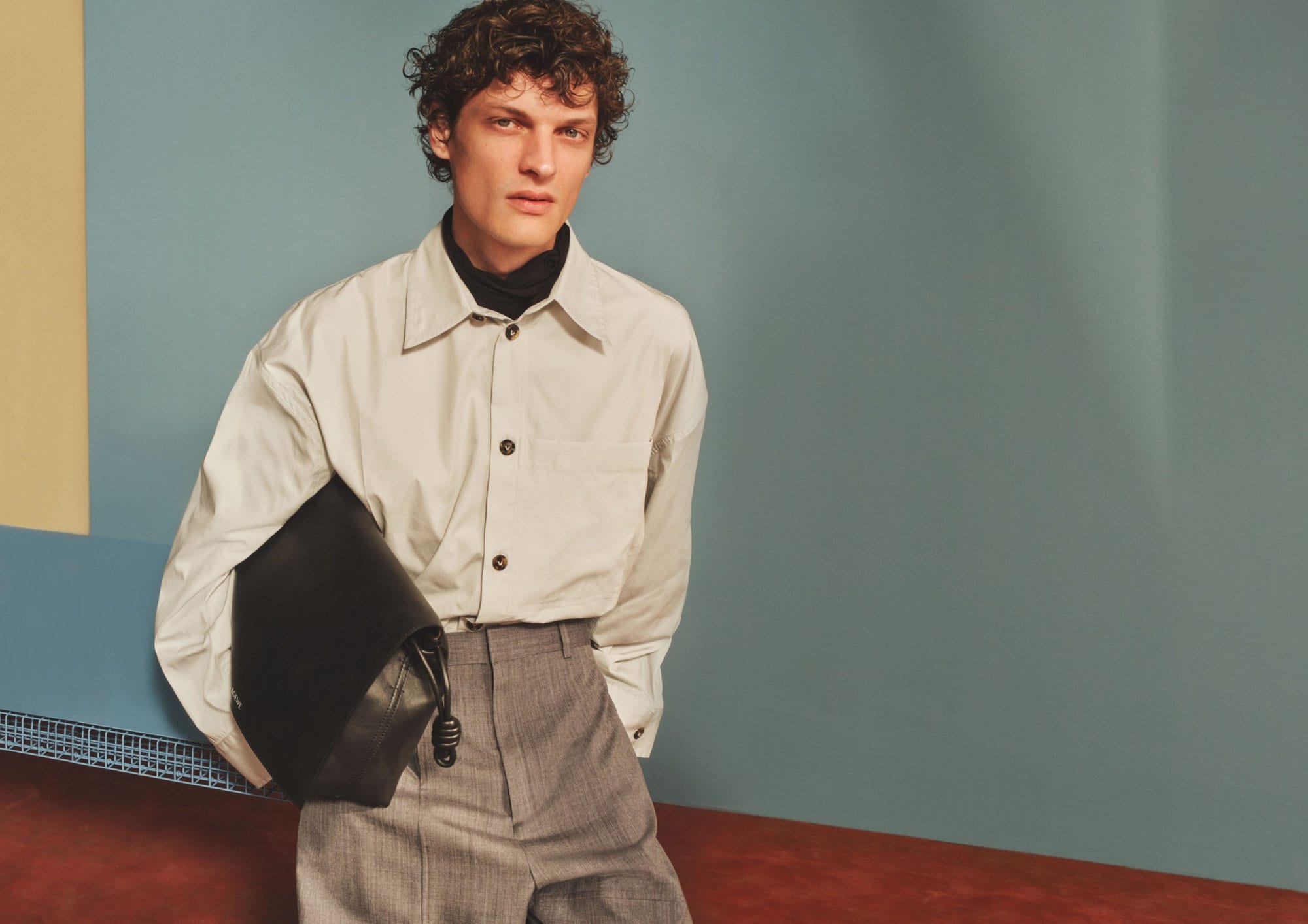 Male model with curly dark hair wearing an off-white button-down shirt and grey trousers holding a black leather bag