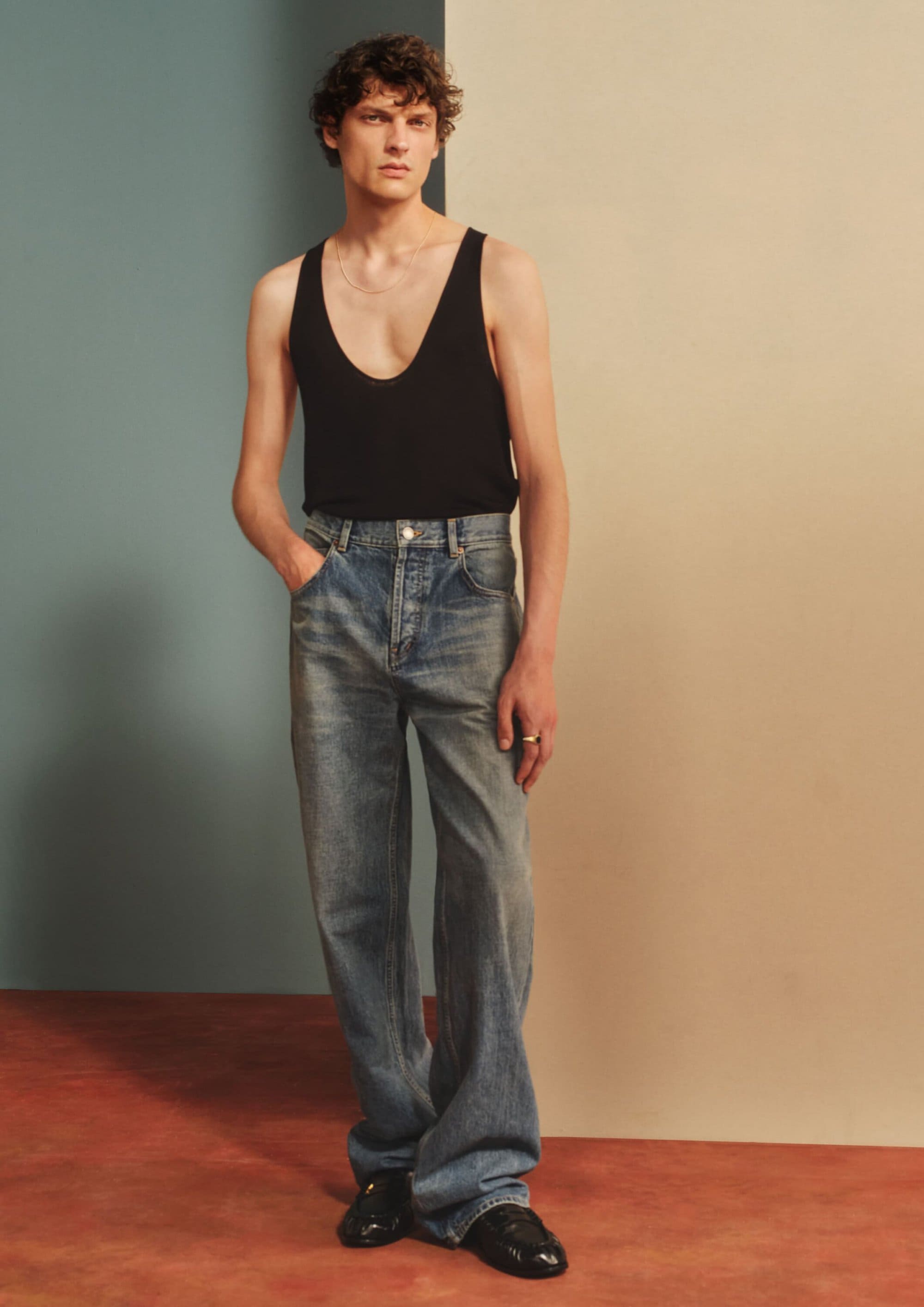 A young male model in a black tank top and blue denim jeans standing against a multi-toned studio background.