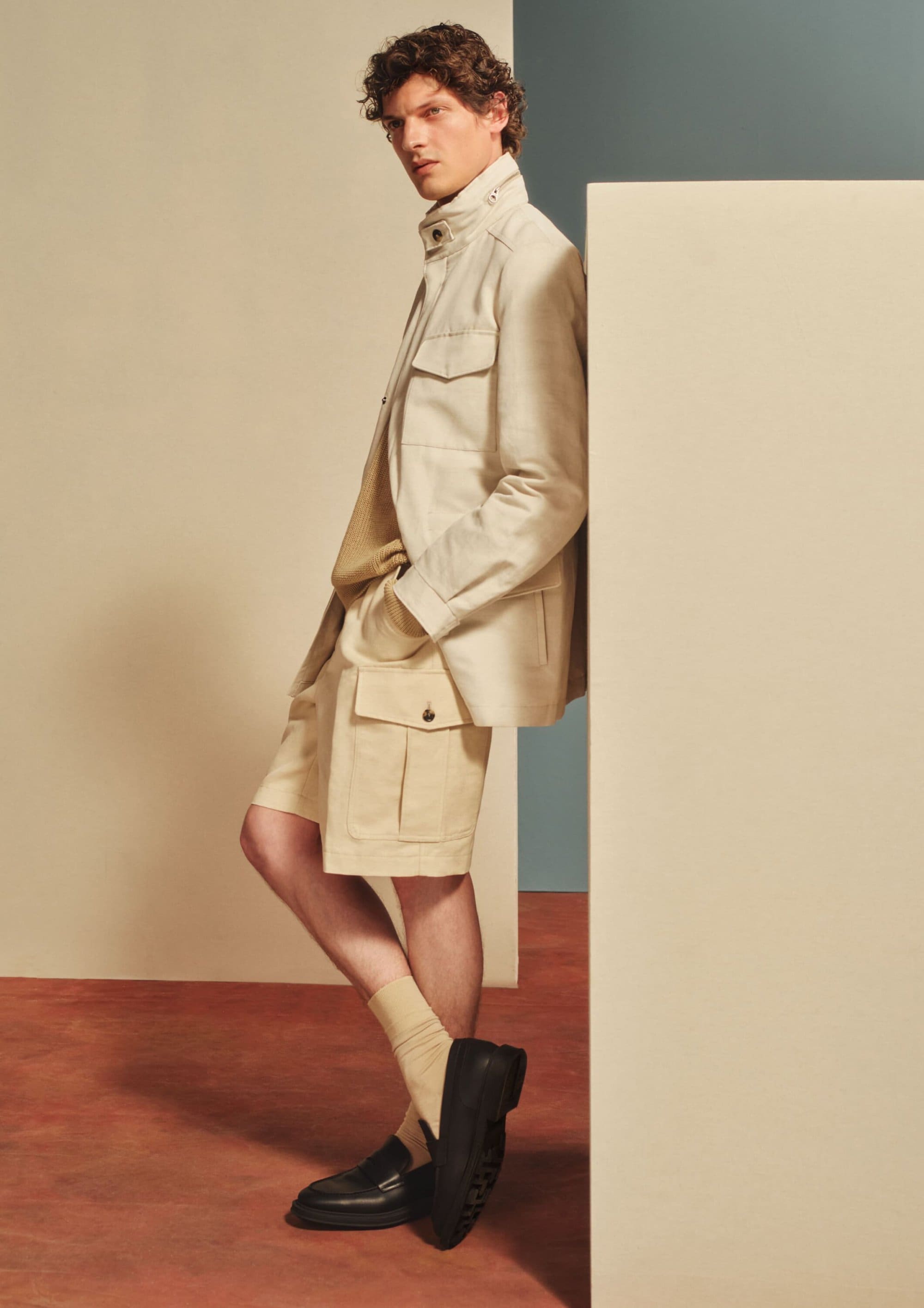 Male model wearing cream utility jacket, shorts and black loafers leaning against a pale geometric studio backdrop