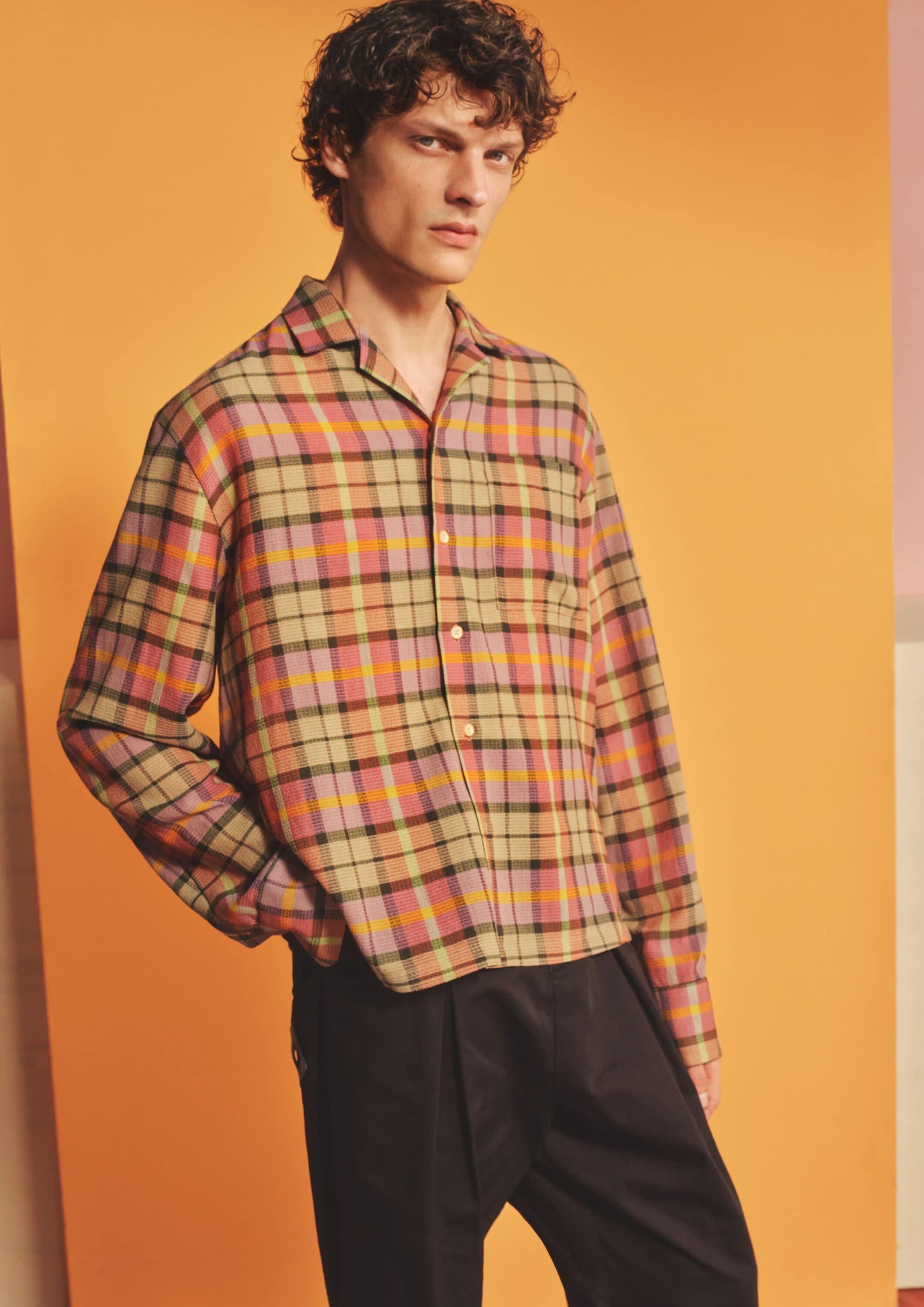 Male model wearing a multi-coloured plaid button-down shirt and dark trousers against an orange studio backdrop.