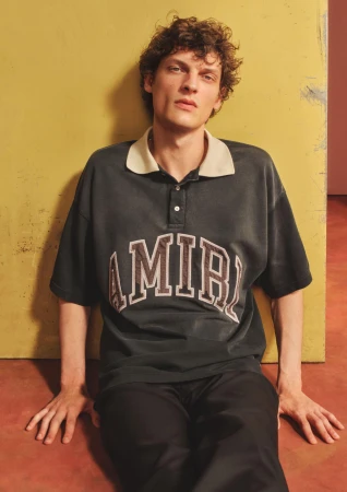Young man with curly brown hair wearing a dark polo shirt leaning against a yellow wall for a Mr Porter campaign.