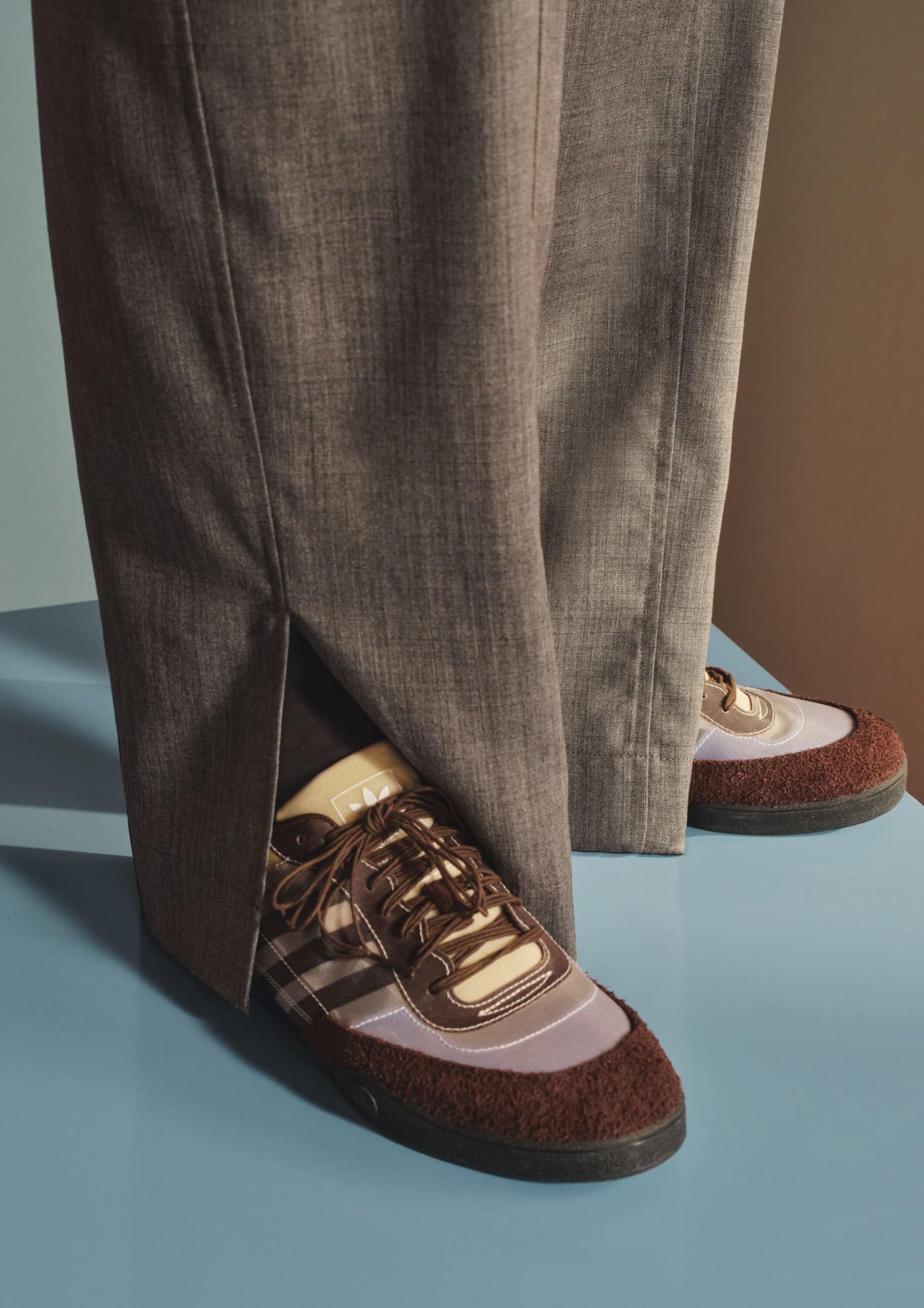 Close up of brown and cream sneakers worn with grey pleated trousers standing on a blue surface for Mr Porter.
