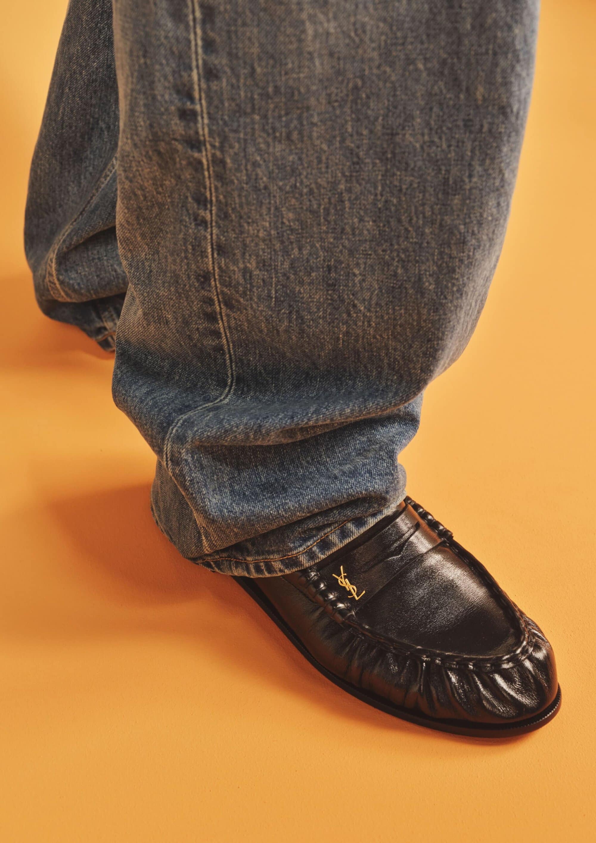 Close-up of a person wearing baggy blue denim jeans and black leather loafers against an orange studio background.