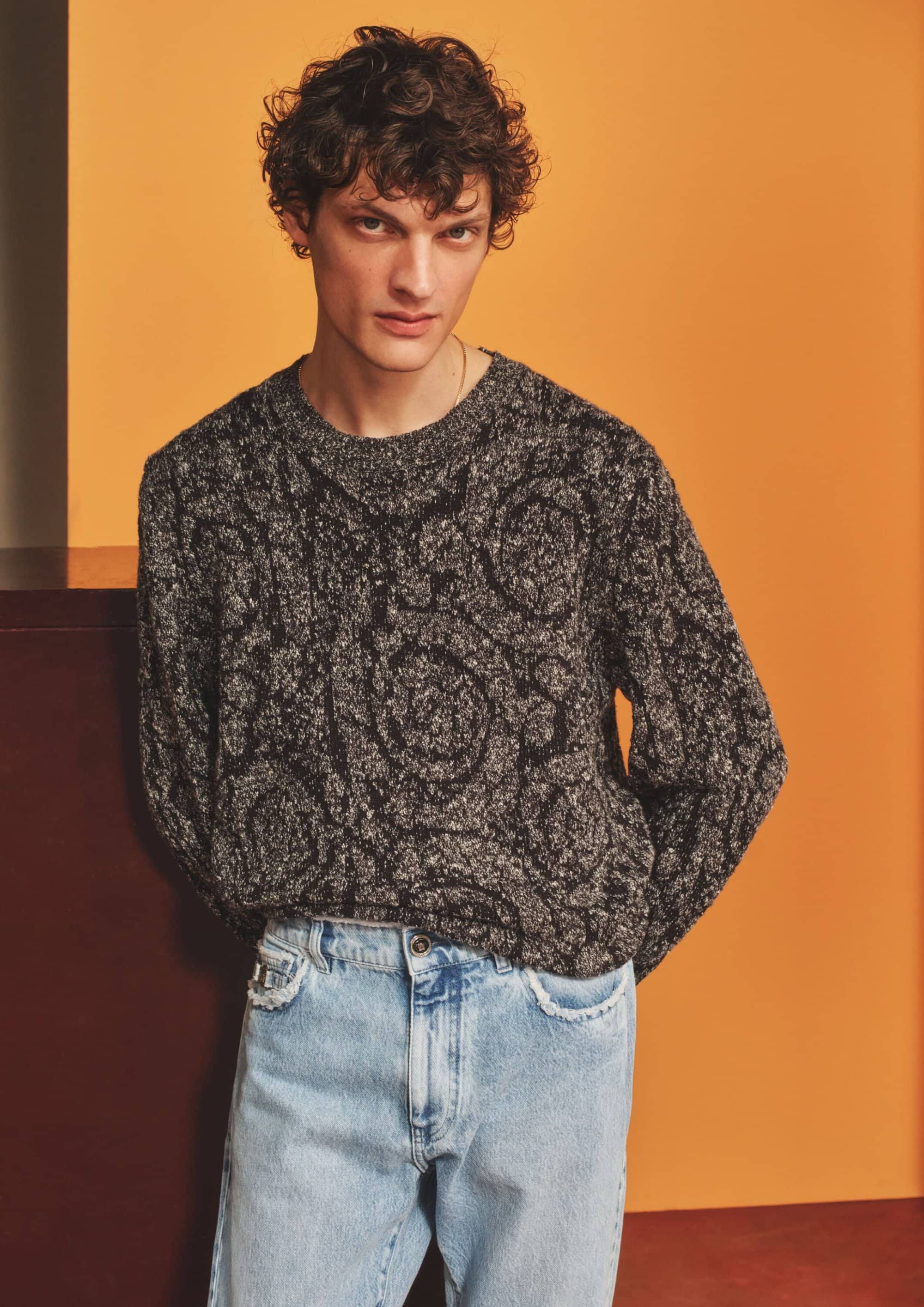A young male model with dark curly hair wearing a patterned dark grey sweater and light blue jeans against a warm studio backdrop.