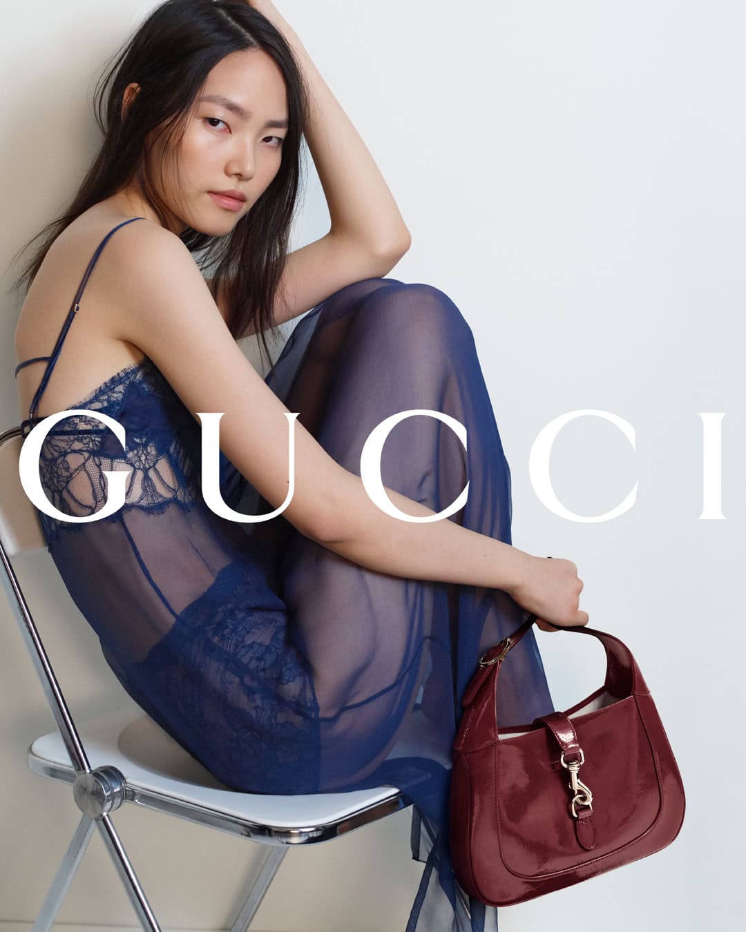Model seated on a white chair in a blue sheer lace slip dress holding a burgundy handbag for Gucci Fall 2024 campaign.