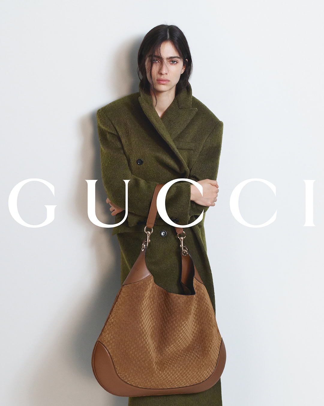 Model in olive green coat holding brown leather shoulder bag against white wall for Gucci Fall 2024 campaign photographed by David Sims