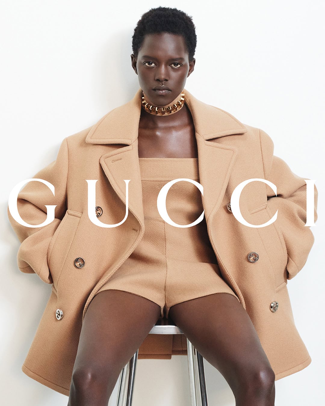 Model wearing a camel coat and choker necklace seated on a stool against a plain white background for the Gucci Fall 2024 ad campaign.