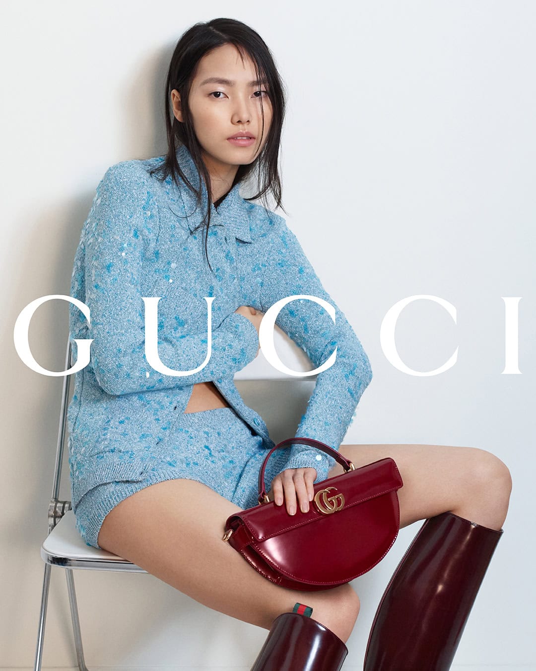 Model seated on a white chair wearing a blue textured top and shorts, holding a dark red patent handbag against a plain background