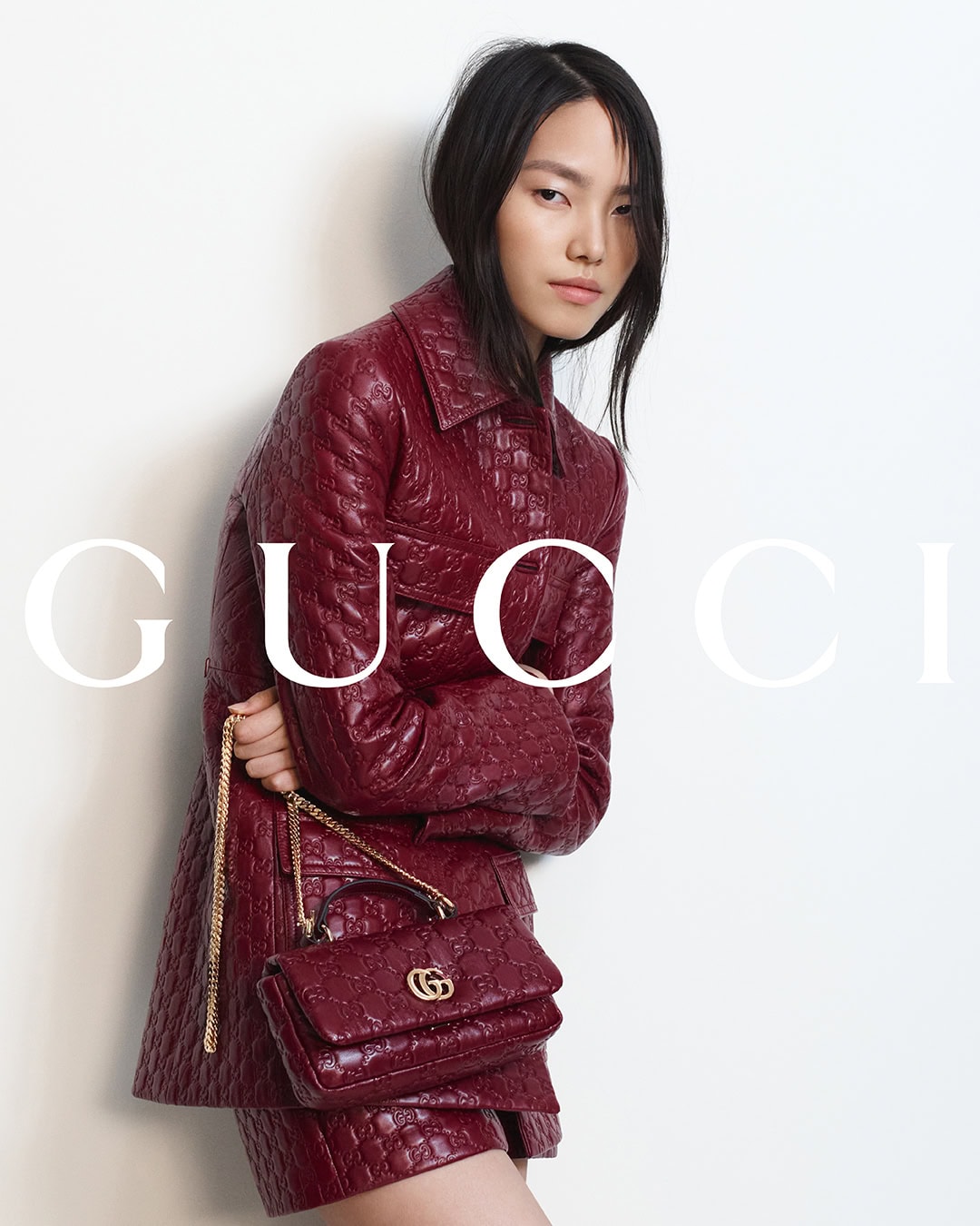 Model in embossed burgundy leather jacket holding matching handbag against plain white studio backdrop