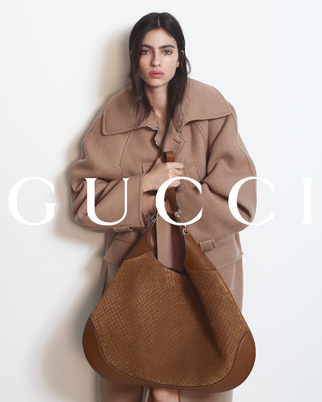 Model in oversized brown coat holding a large brown leather shoulder bag against a plain white background with Gucci typography overlay