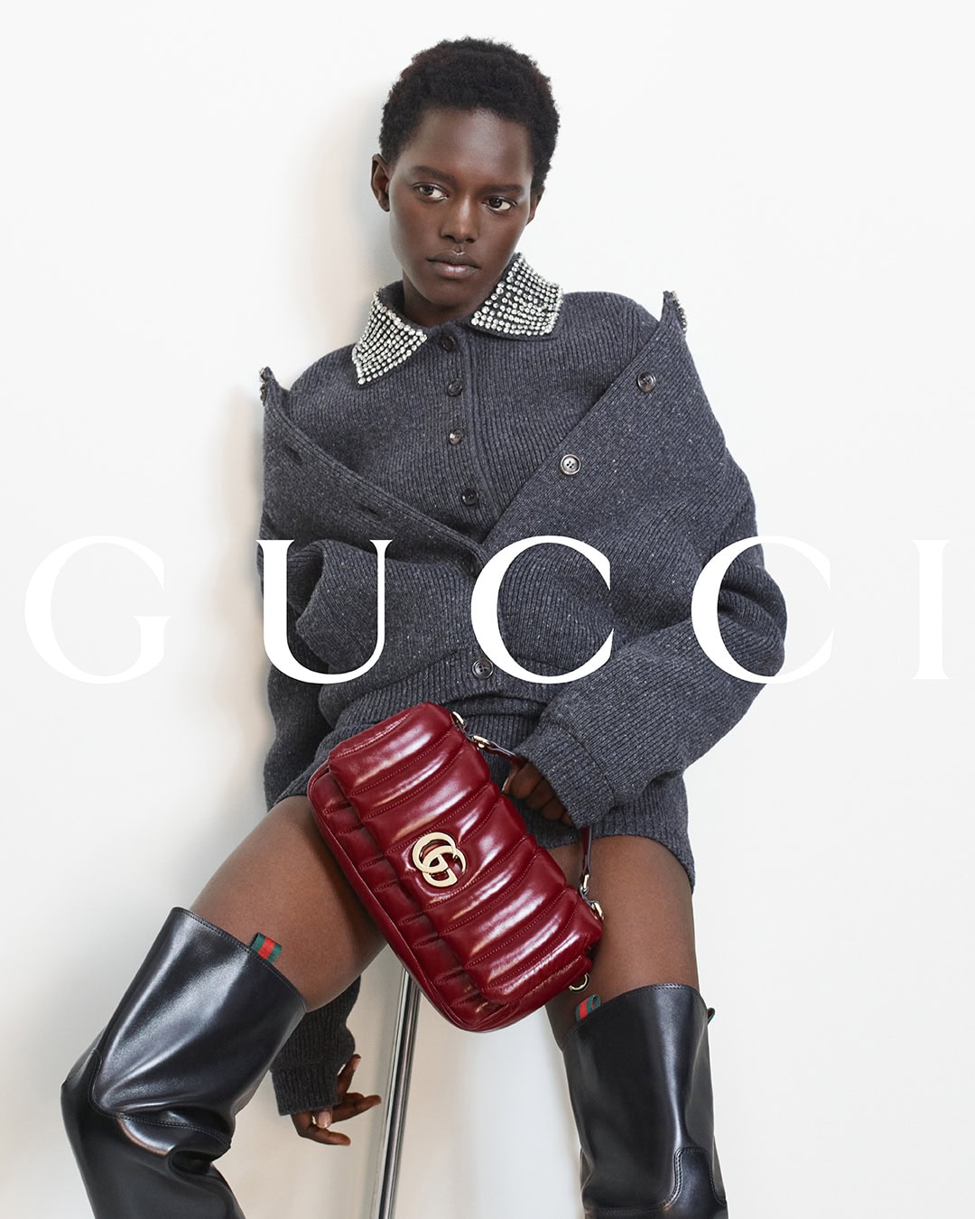 Model wearing a grey embellished cardigan and black boots holds a quilted red handbag against a white background