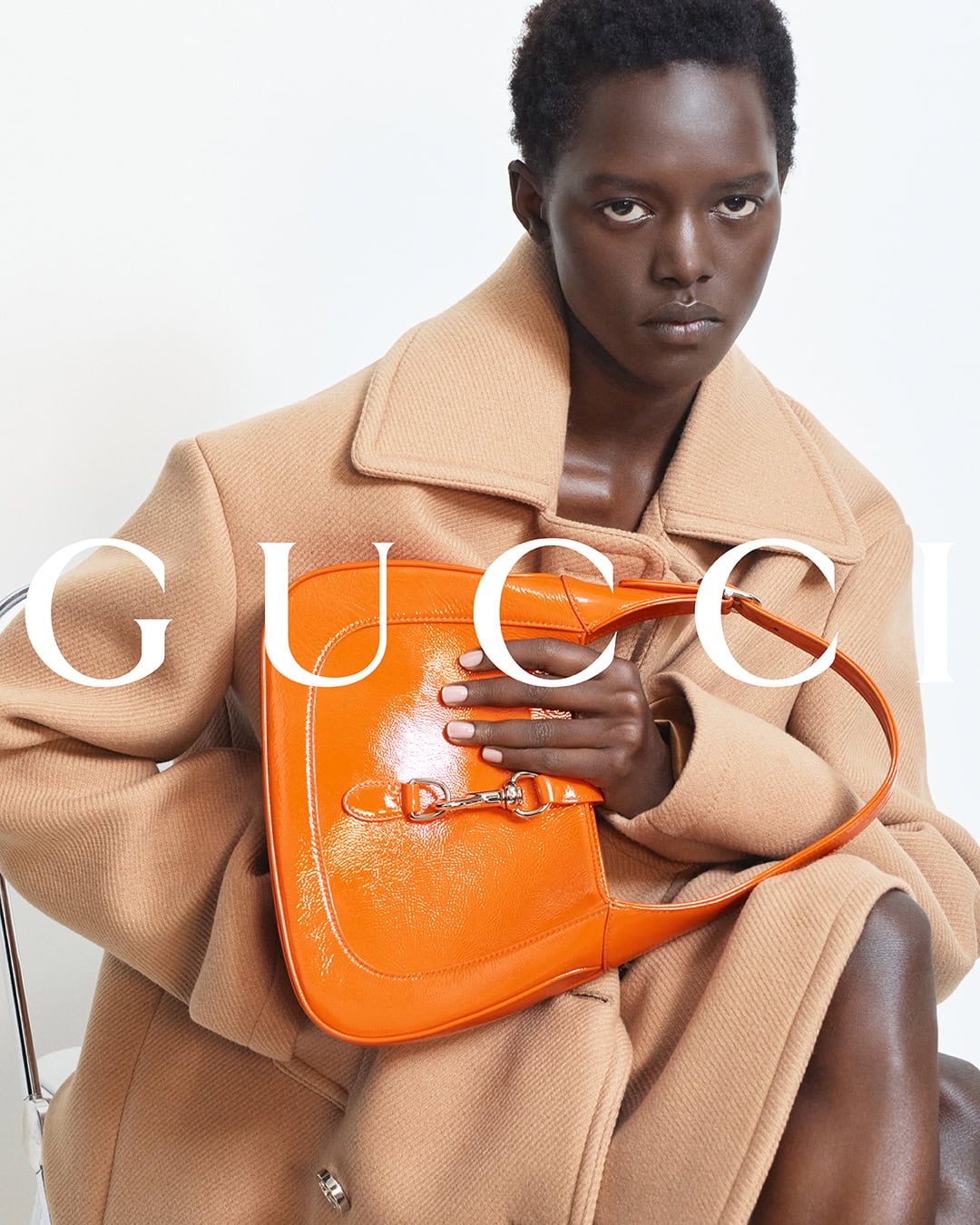 Model wearing a camel coat holding a bright orange patent leather shoulder bag against a white background