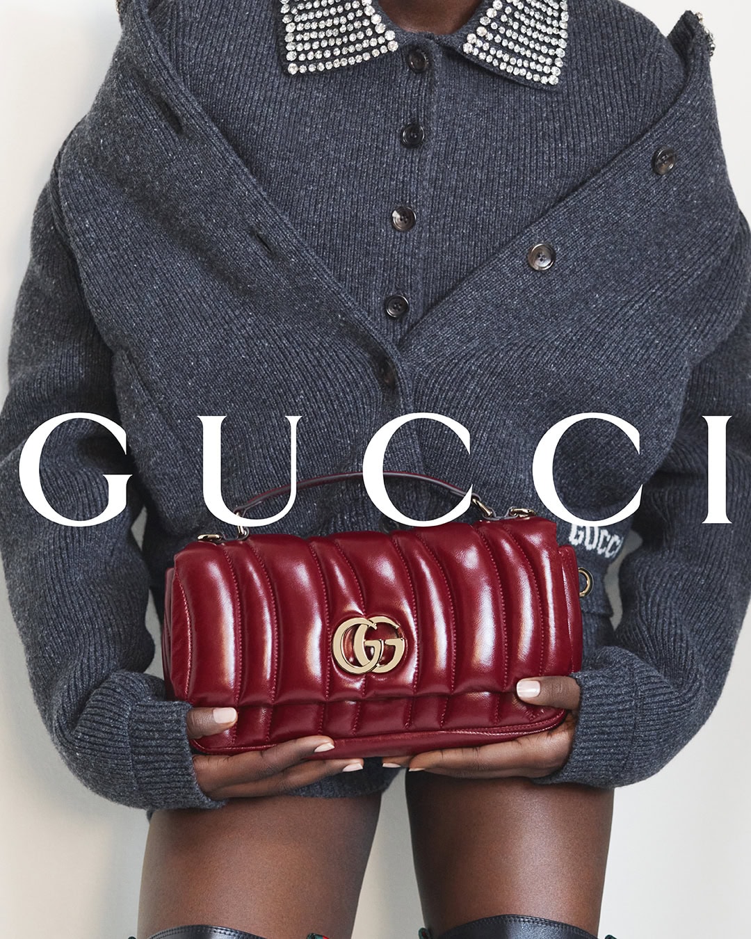 Model holding a quilted red leather handbag wearing a grey cardigan with crystal collar for Gucci Fall 2024 campaign