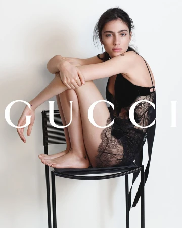 Model wearing a black lace slip dress seated on a modern black wire chair against a white wall.