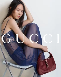 Model seated on a white chair in a blue sheer lace slip dress holding a burgundy handbag for Gucci Fall 2024 campaign.