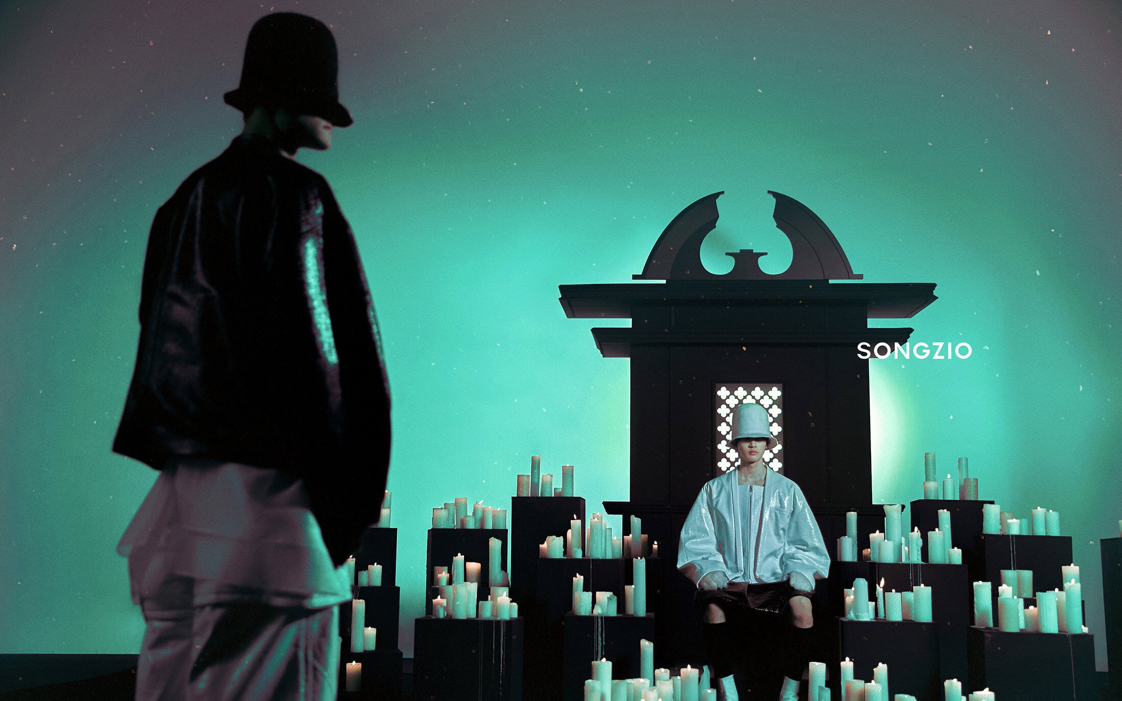 Two models in dark attire and hats on a green-lit stage surrounded by numerous burning candles for Songzio Fall 2024 campaign