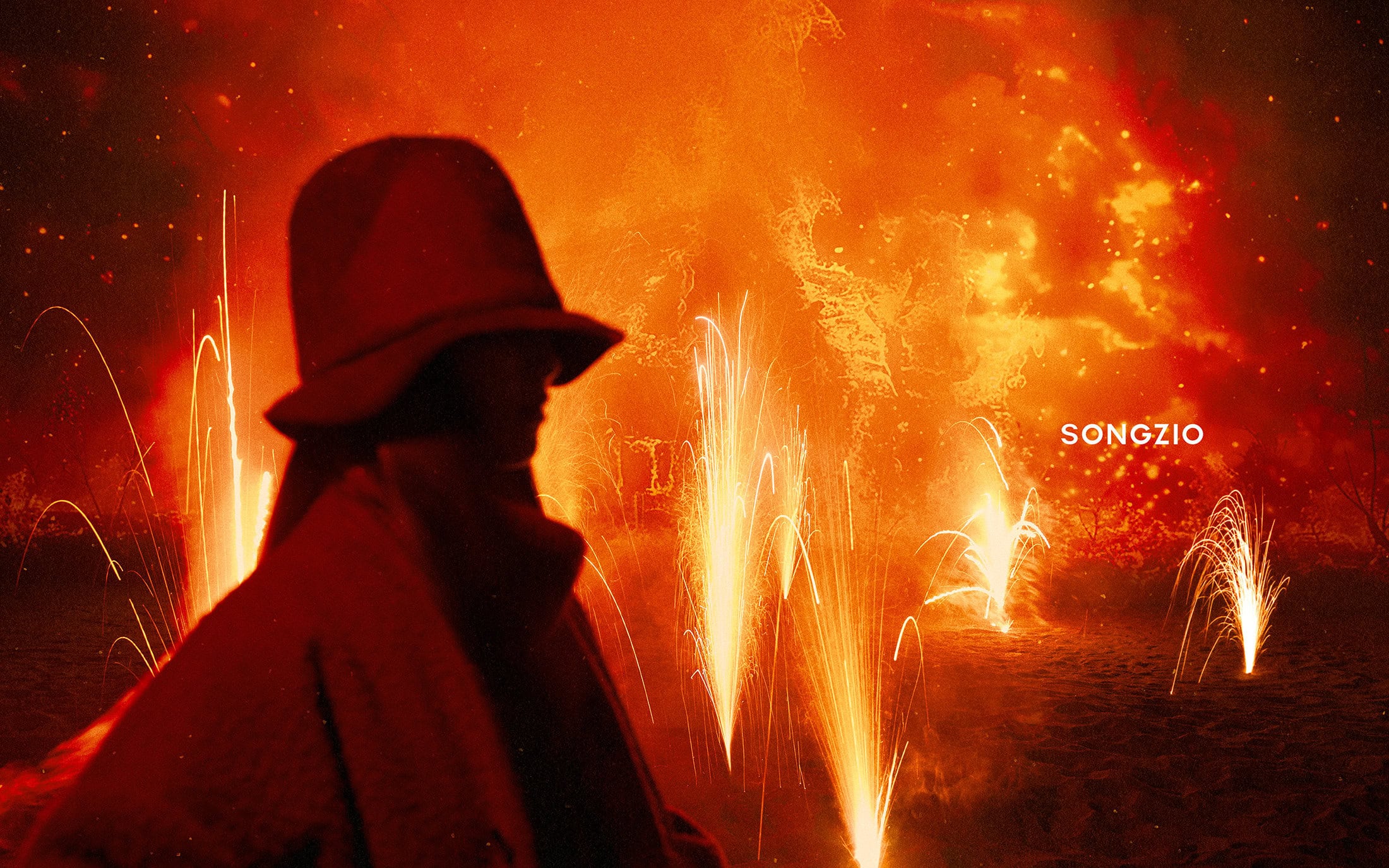Silhouette of a person wearing a hat and coat against an intense fiery background with glowing orange sparks and firework fountains.