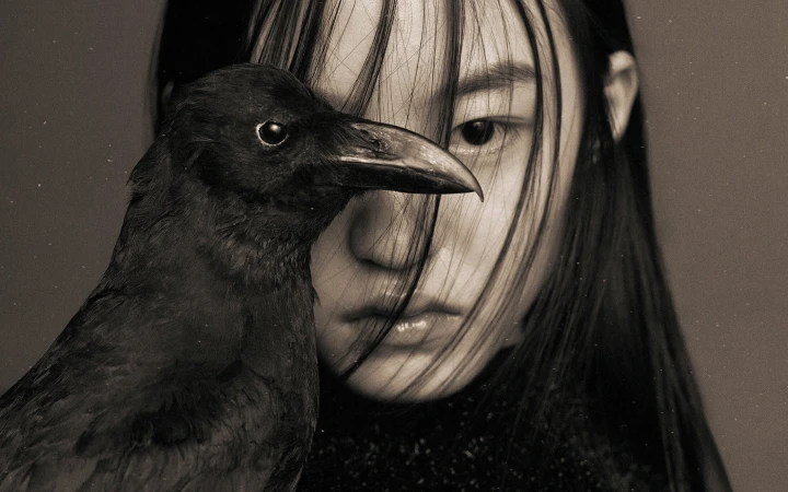 Close-up portrait of a model with dark hair partially obscuring her face, next to a black crow in the foreground.