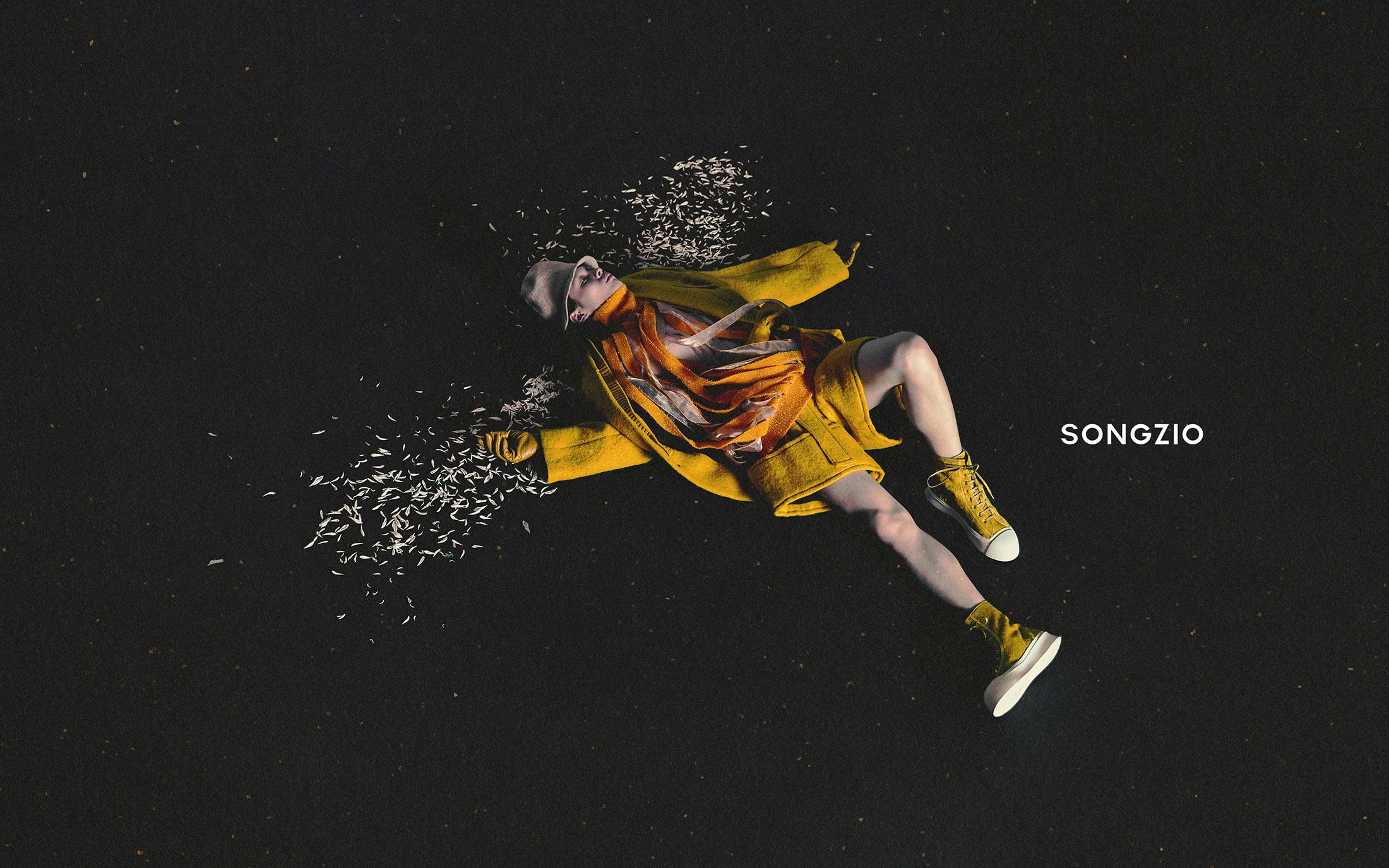 Model lying on a dark textured background wearing a vibrant yellow coat and matching yellow high-top sneakers surrounded by scattered petals.
