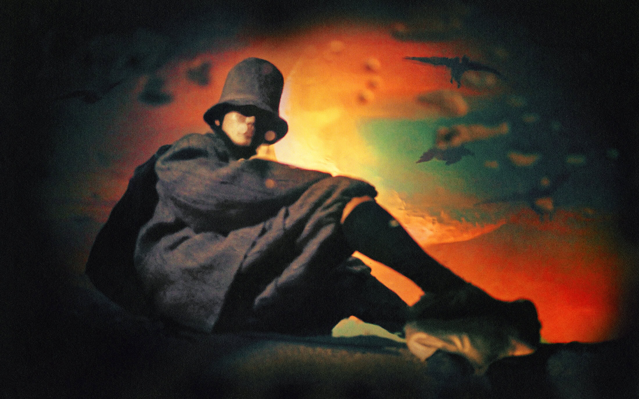 Seated figure wearing a dark wide-brimmed hat and layered coat against a colorful, fiery sky backdrop.
