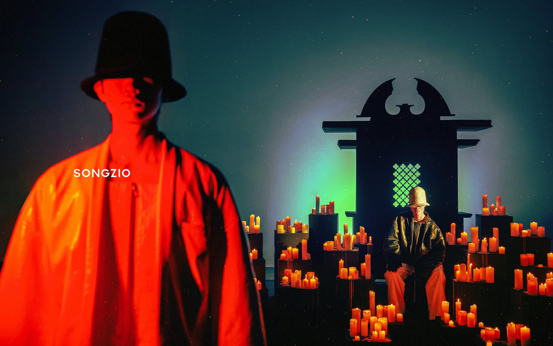 Model in red lighting with black hat in the foreground and a second seated model surrounded by candles against a dark background