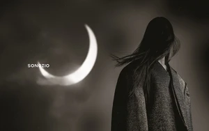 Monochrome portrait of a person with long dark hair wearing a dark jacket, facing away against a large glowing crescent moon background.