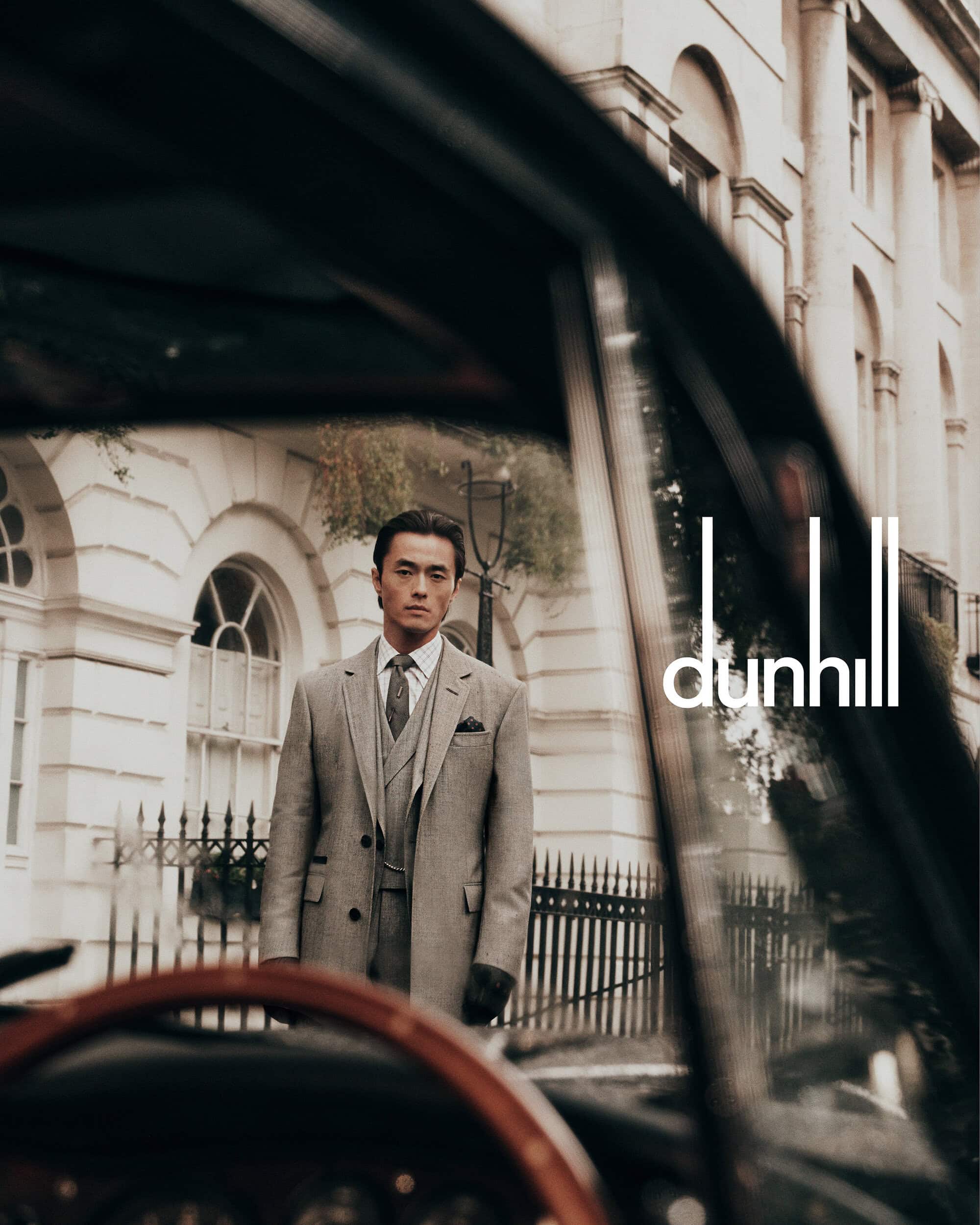 Man wearing a grey three-piece suit standing outside a classical building, framed through a car windshield.