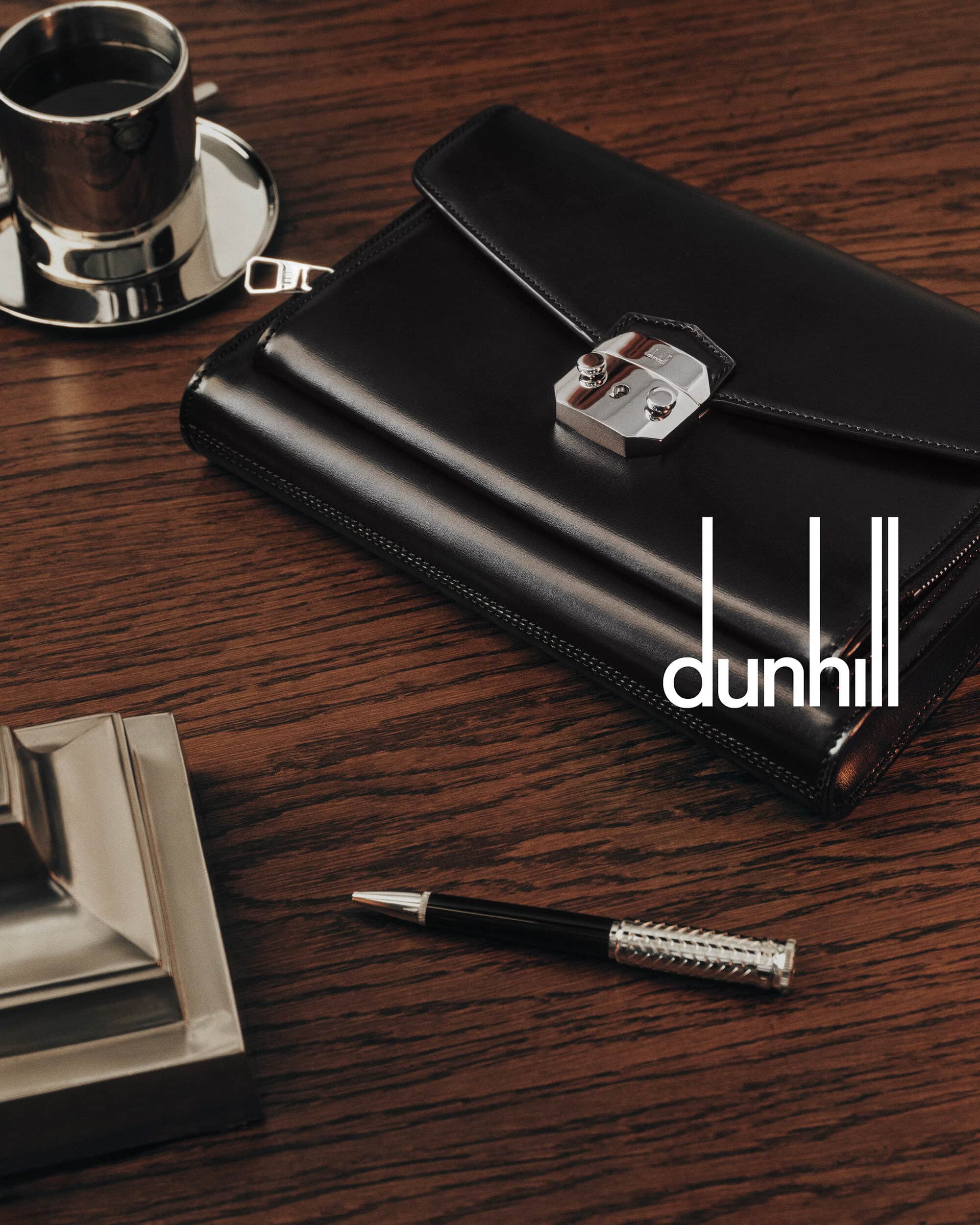 Black leather document case with silver clasp resting on a dark wood grain table alongside a silver cup and pen.