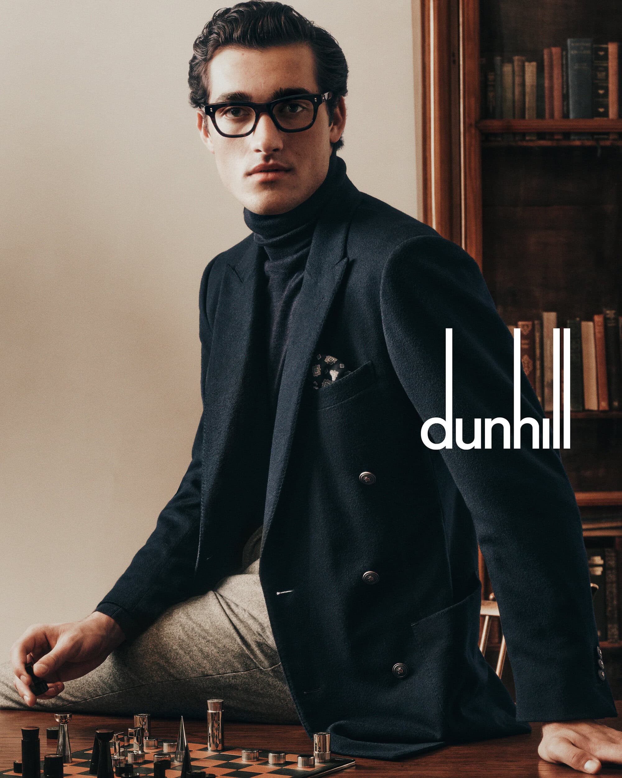 A man wearing glasses and a dark blazer plays chess in a library setting for Dunhill.