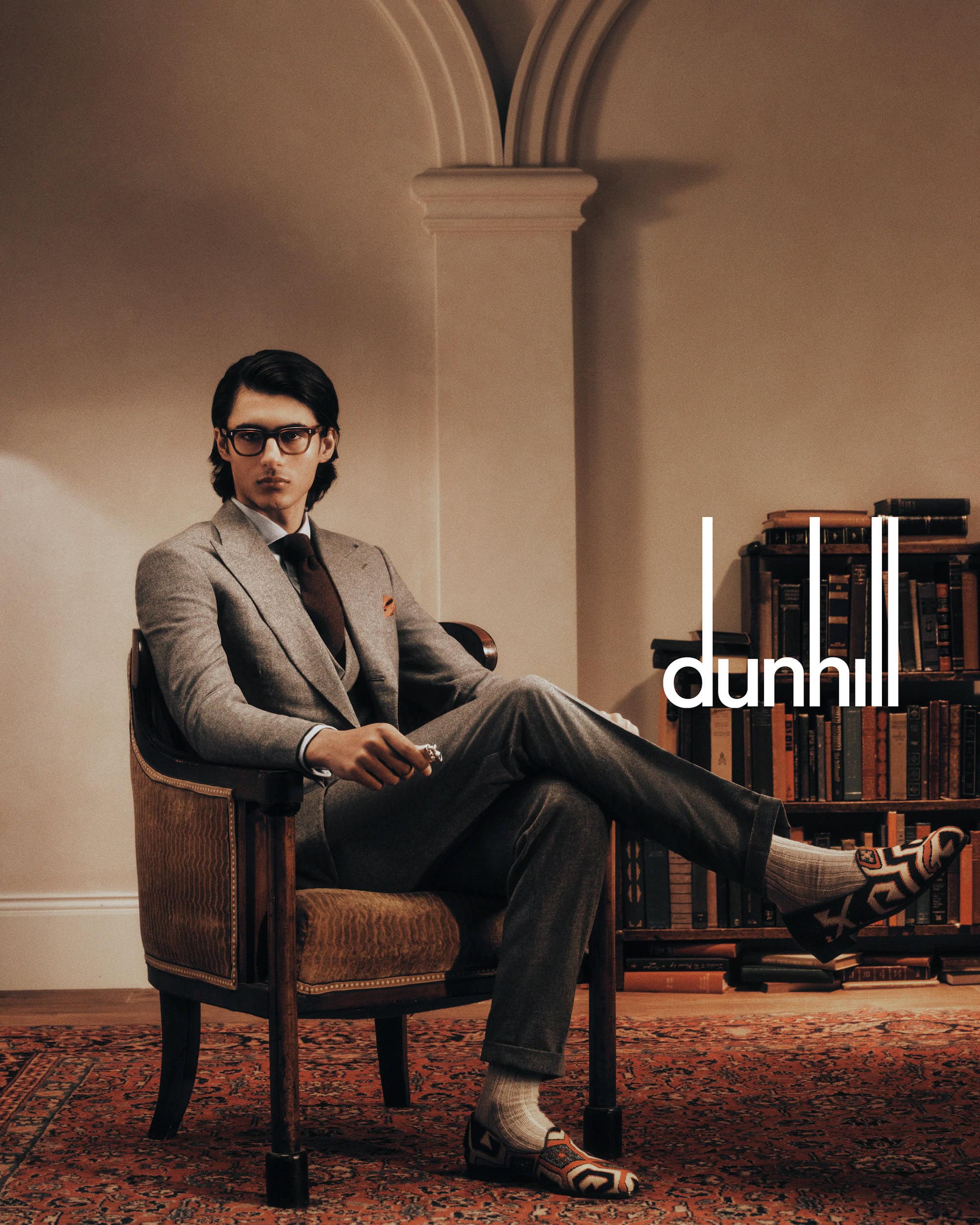 Man wearing a grey suit and glasses seated in an armchair inside a library for Dunhill Fall 2024 campaign