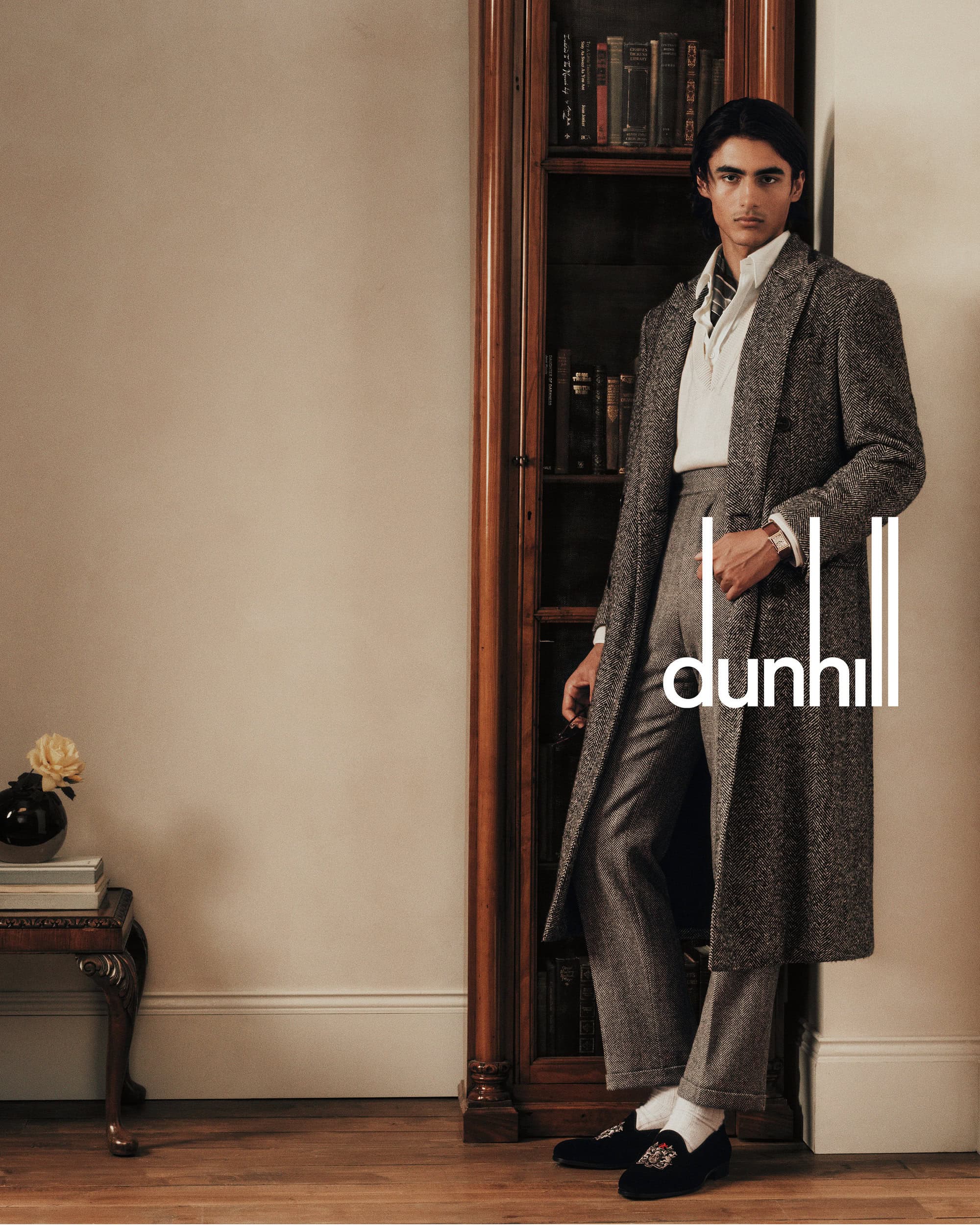 A man in a grey tweed coat standing beside a wooden bookcase for Dunhill Fall 2024 Chapter 2 ad campaign.