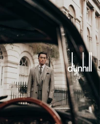 Man wearing a grey three-piece suit standing outside a classical building, framed through a car windshield.