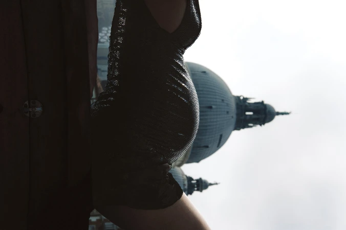 Close-up of a model wearing a dark metallic chainmail dress outdoors against an architectural dome and pale sky.