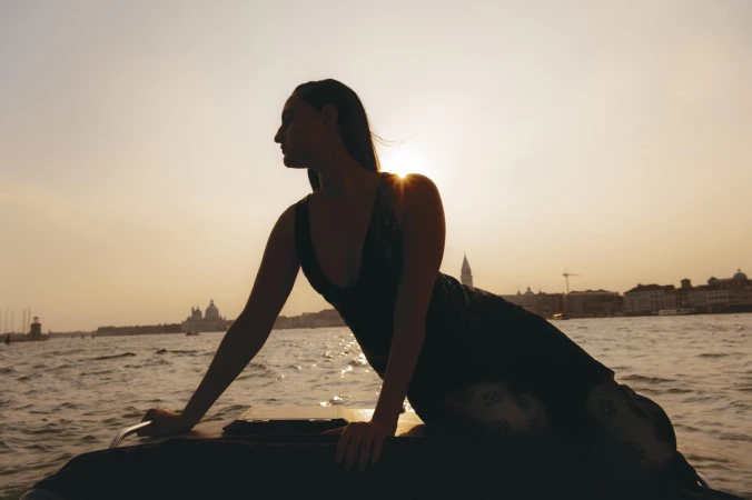 Model posed on a boat against a sunset cityscape with silhouetted water and sky