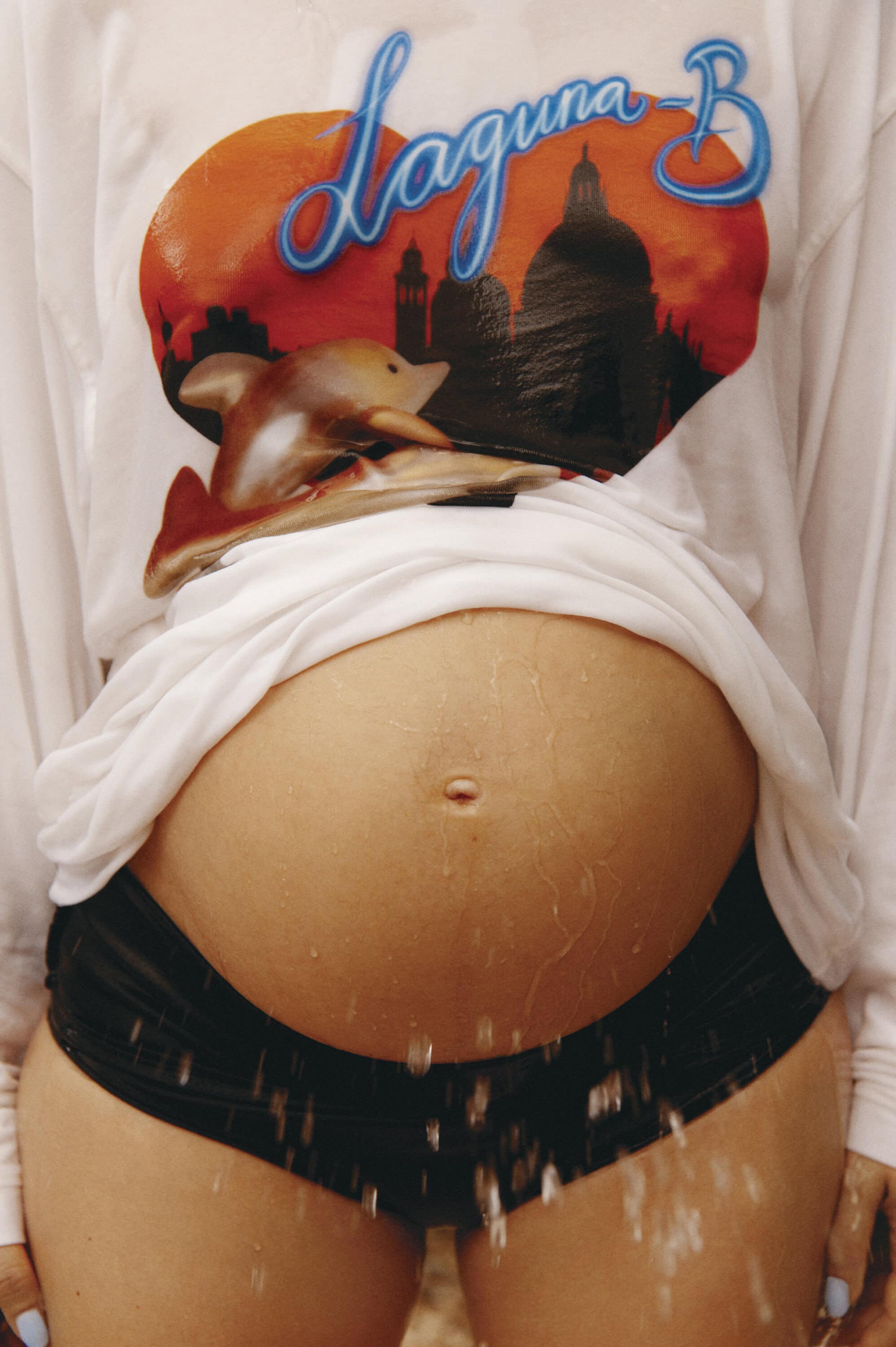 Pregnant woman wearing white graphic t-shirt and dark shorts with water droplets falling over her body.