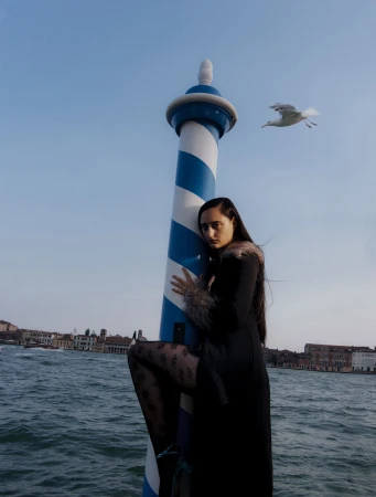Model hugging a blue and white striped mooring pole by the water in Venice, Italy with a flying seagull in the background.