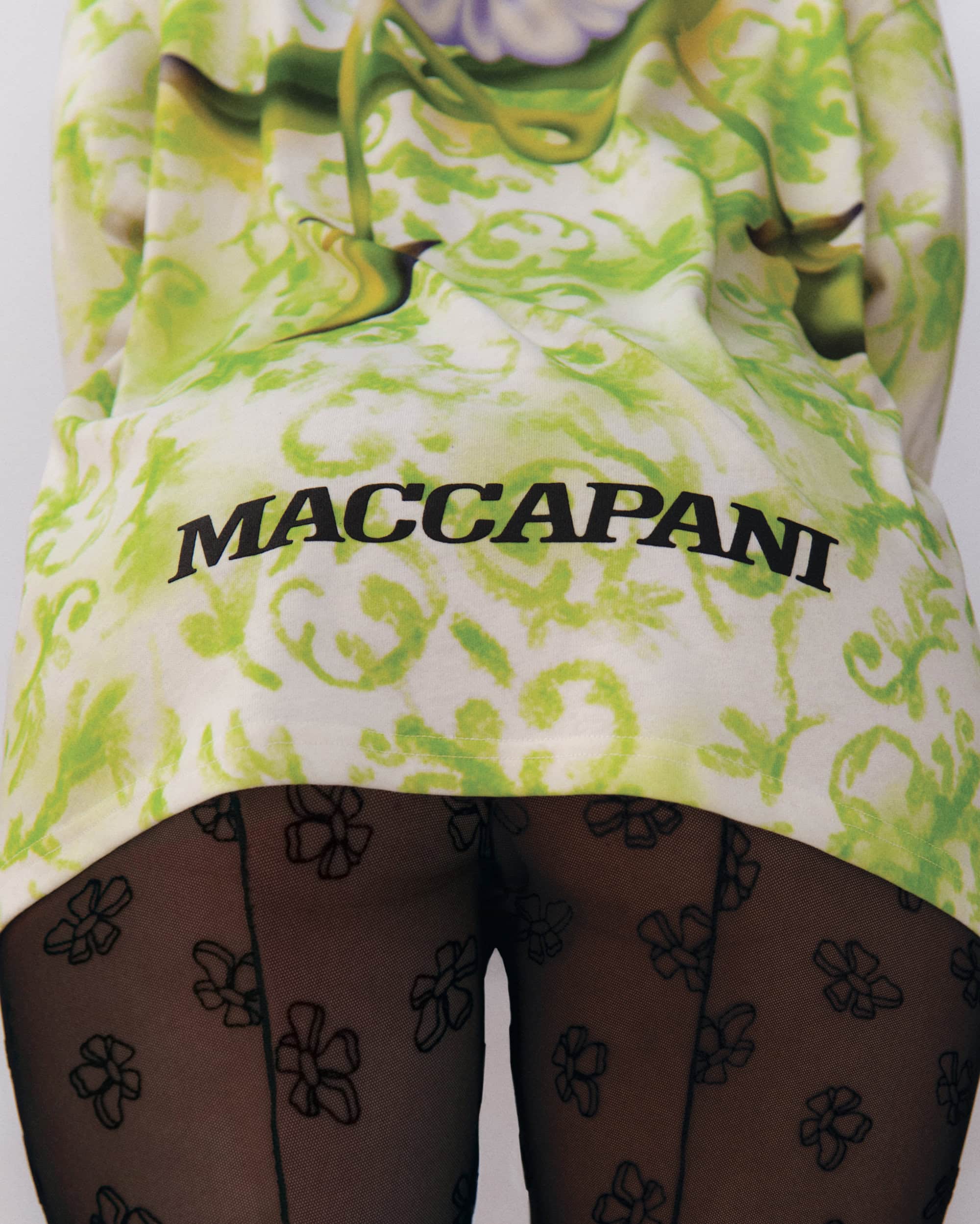 Model wearing patterned green top with Maccapani logo and sheer black patterned tights against white background.