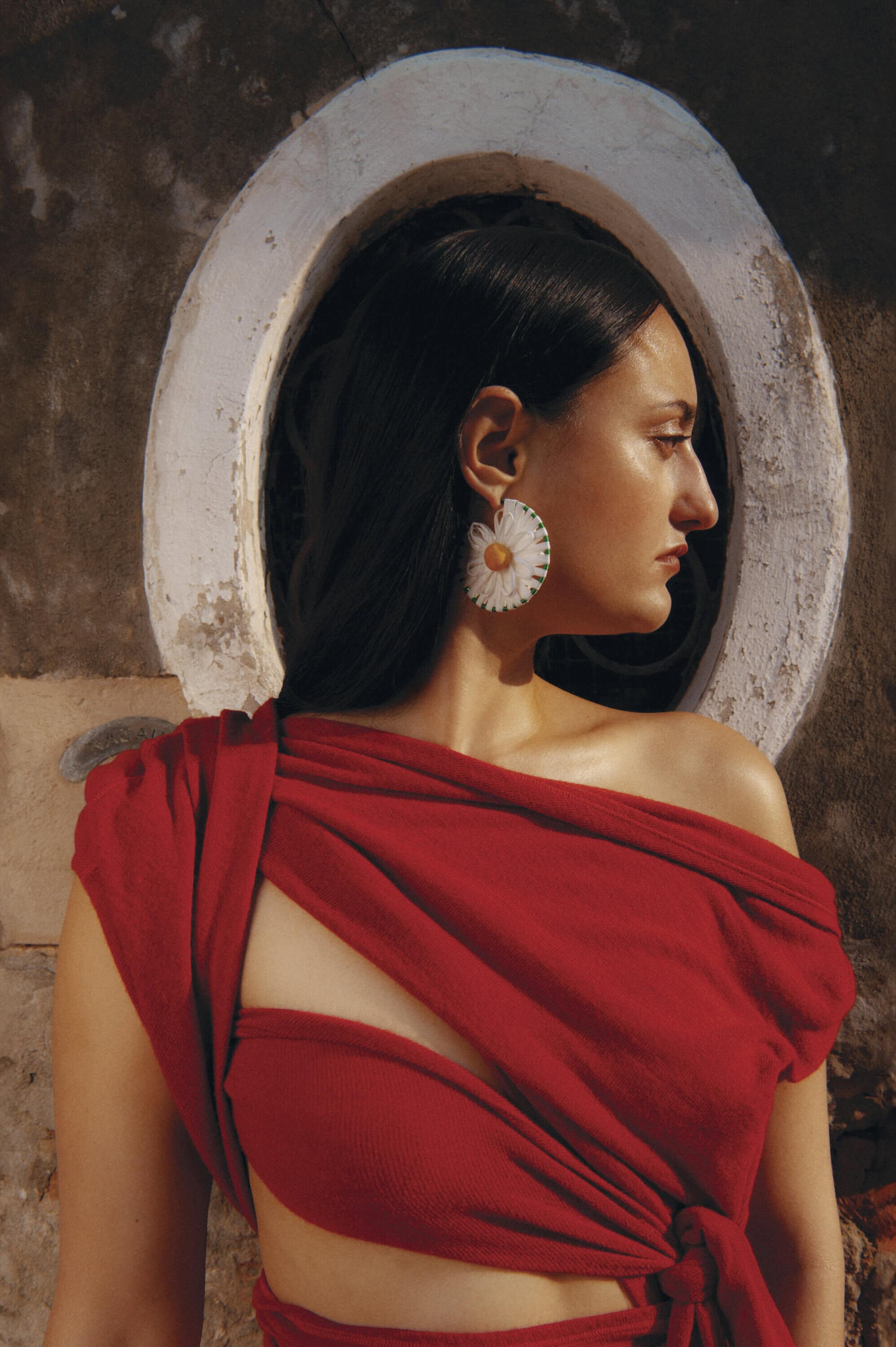 Model in red outfit wearing large daisy earring posing against a textured wall with an arched architectural opening.
