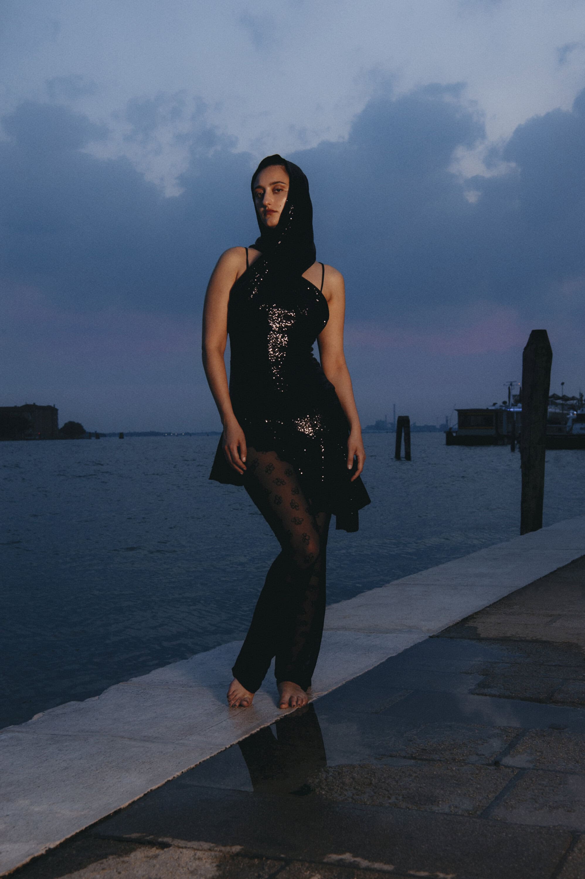 Model standing on a stone pier by the water at dusk wearing a black sequined dress and head covering